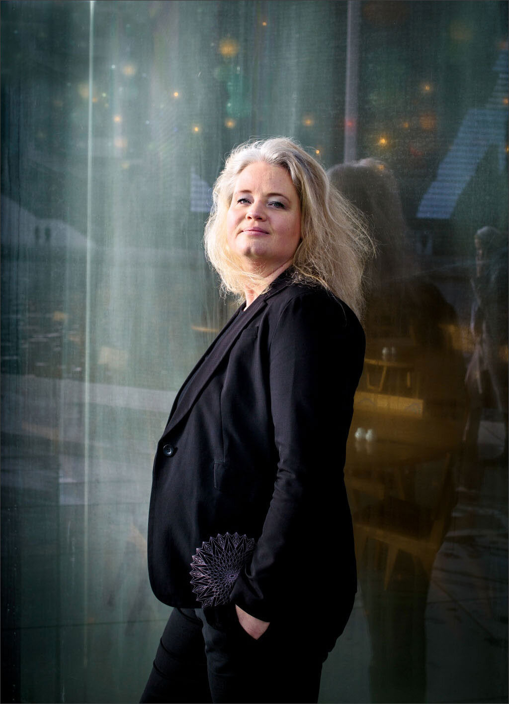 Frau mit blondem, lockigem Haar, schwarze Jacke mit Muster am Ärmel, steht vor einer reflektierenden Glaswand.