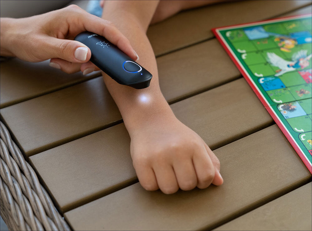 Hand hält Infrarot-Thermometer an Kinderarm, daneben Spielbrett mit bunten Figuren auf Holztisch.