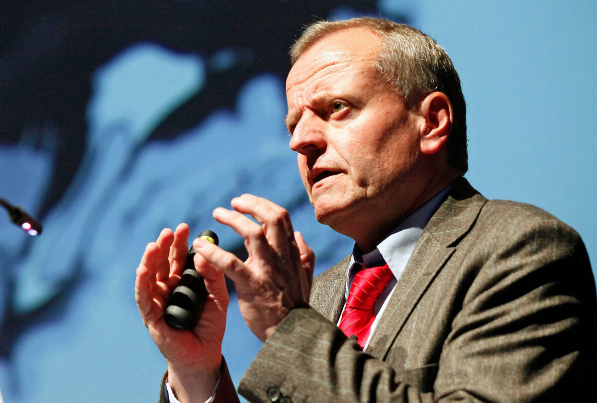 Mann in Anzug mit roter Krawatte hält Mikrofon und spricht vor einer blauen Hintergrundprojektion.