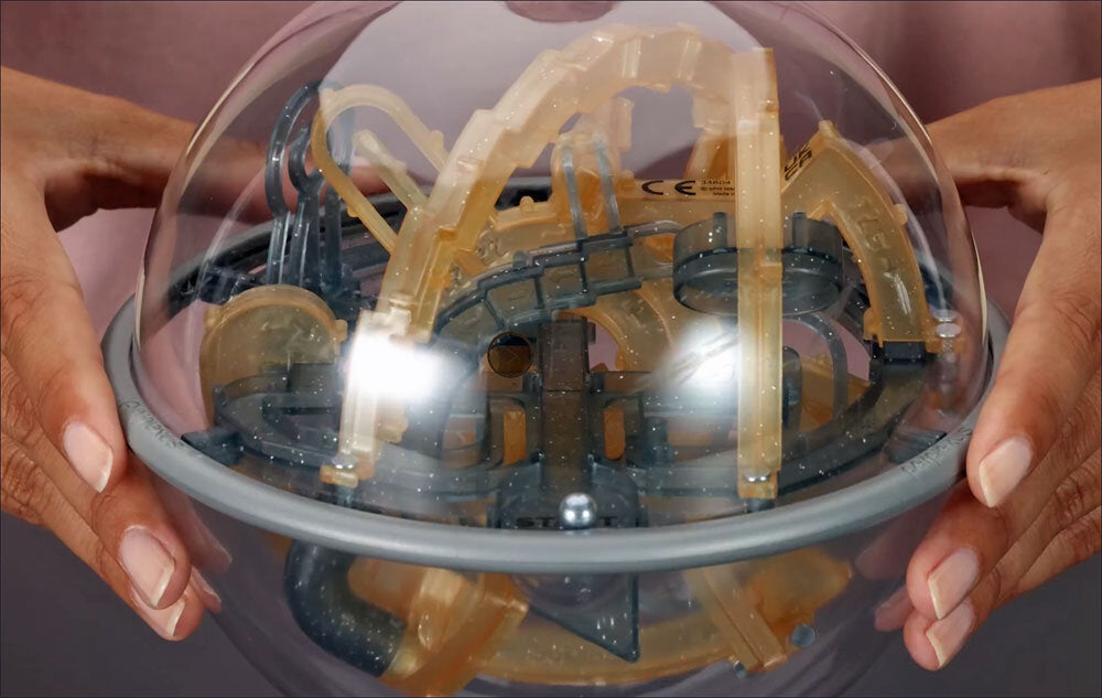 Hand hält transparenten, runden Spielzeug mit gelben und schwarzen Teilen, darunter eine Metallkugel.