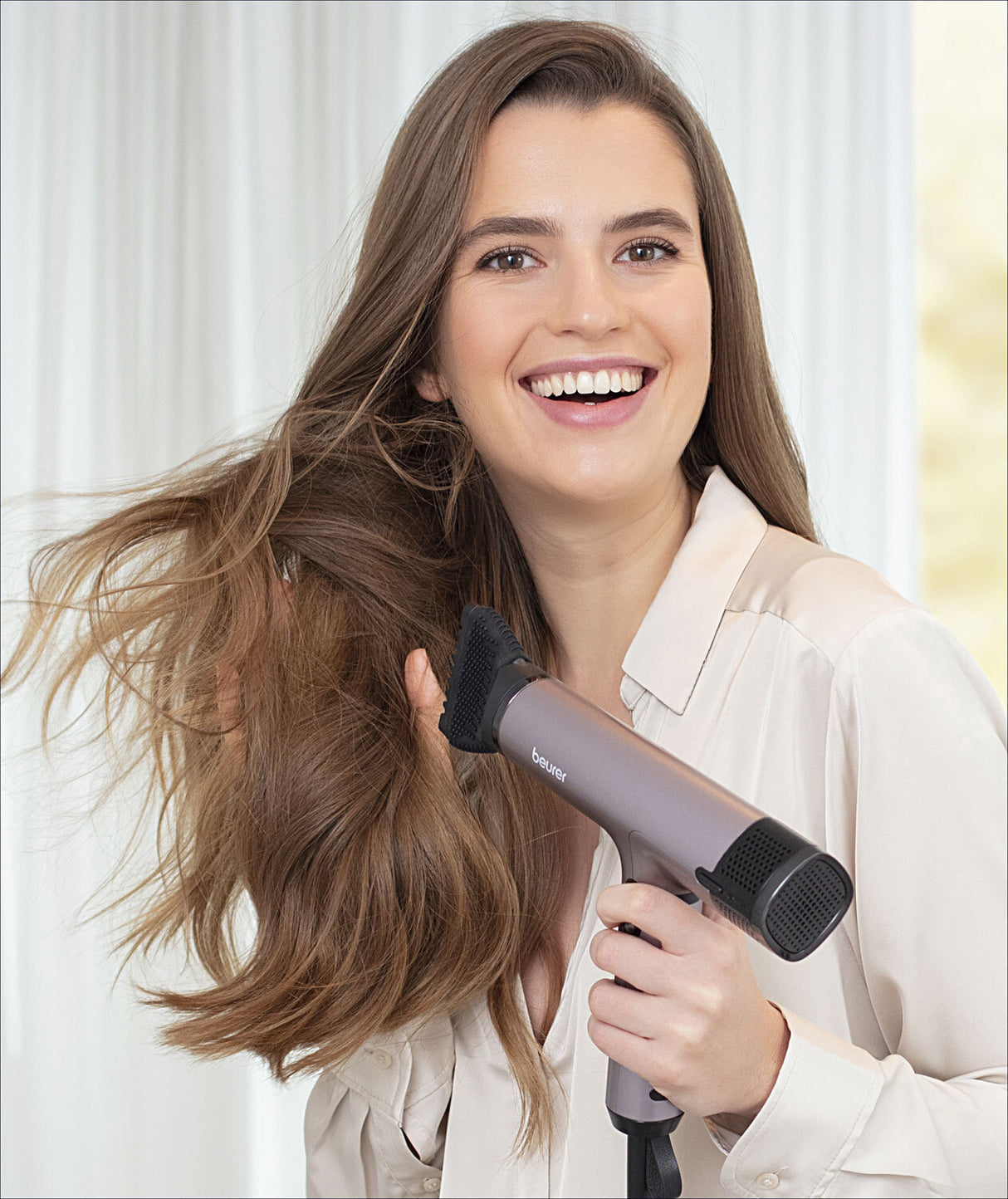 Frau trocknet ihre langen braunen Haare mit einem Haartrockner, lächelt und trägt eine weiße Bluse.