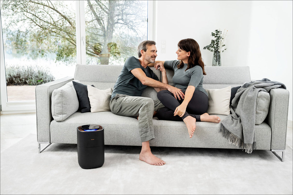 Zwei Menschen sitzen auf einem grauen Sofa, lachen und schauen sich liebevoll an, im Wohnzimmer mit großen Fenstern.