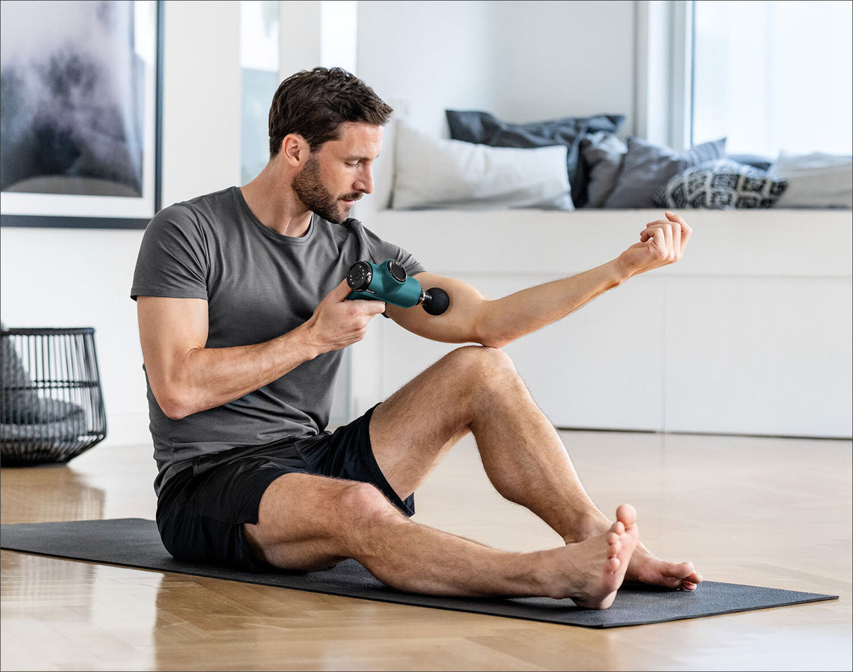 Mann sitzt auf Yogamatte, massiert seinen Arm mit Massagegerät, im Wohnzimmer