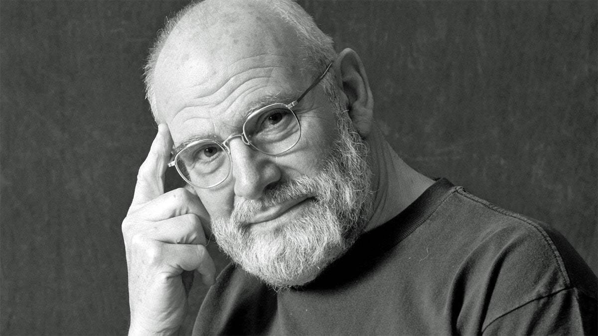 Älterer Mann mit Brille, grauem Bart, trägt schwarzen Pullover, berührt Kopf mit rechter Hand, blickt nach vorne.