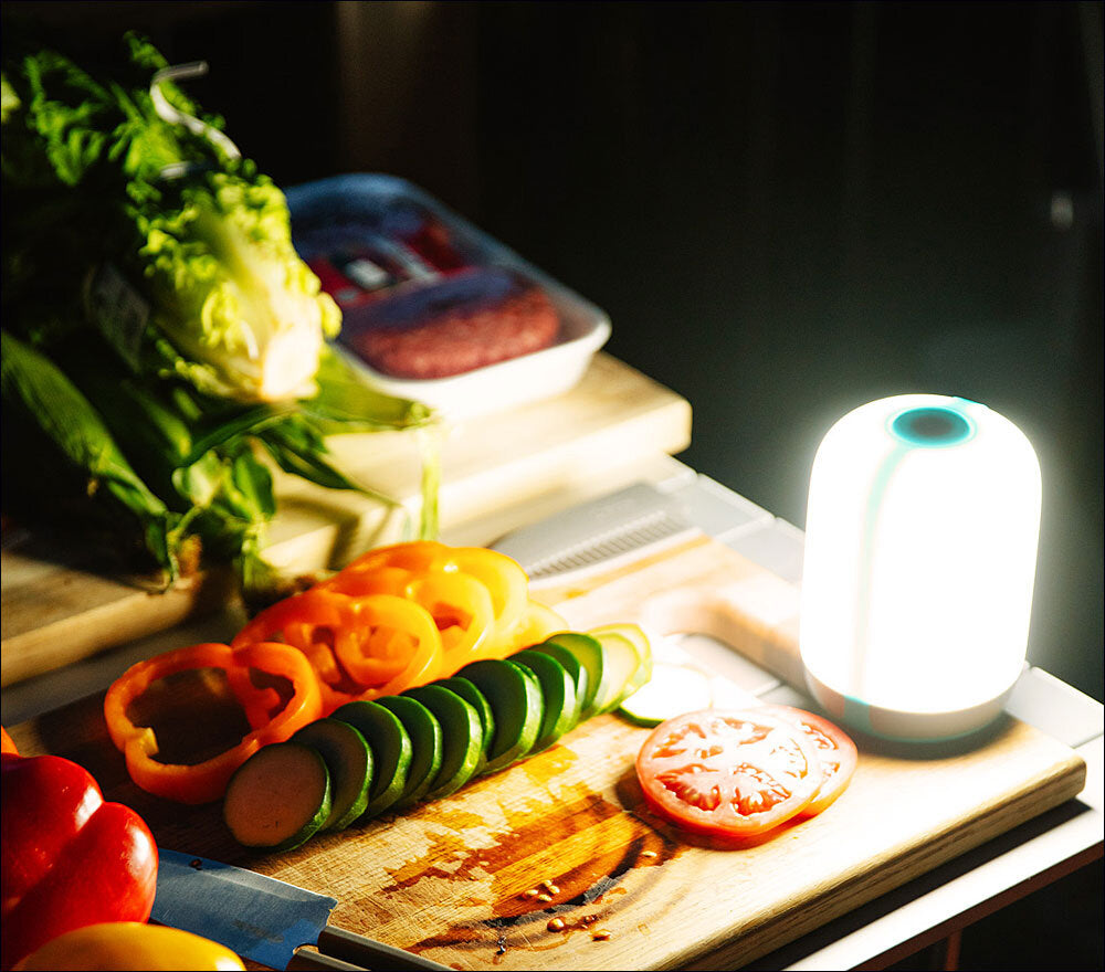Bunte Paprika, Tomate und Zucchini auf Schneidebrett, Küchenlampe leuchtet, im Hintergrund Fleisch und Gemüse.