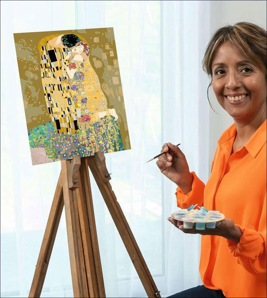 Frau malt ein Bild im Stil von Gustav Klimt auf einer Staffelei, hält einen Pinsel und eine Farbpalette, lächelt.