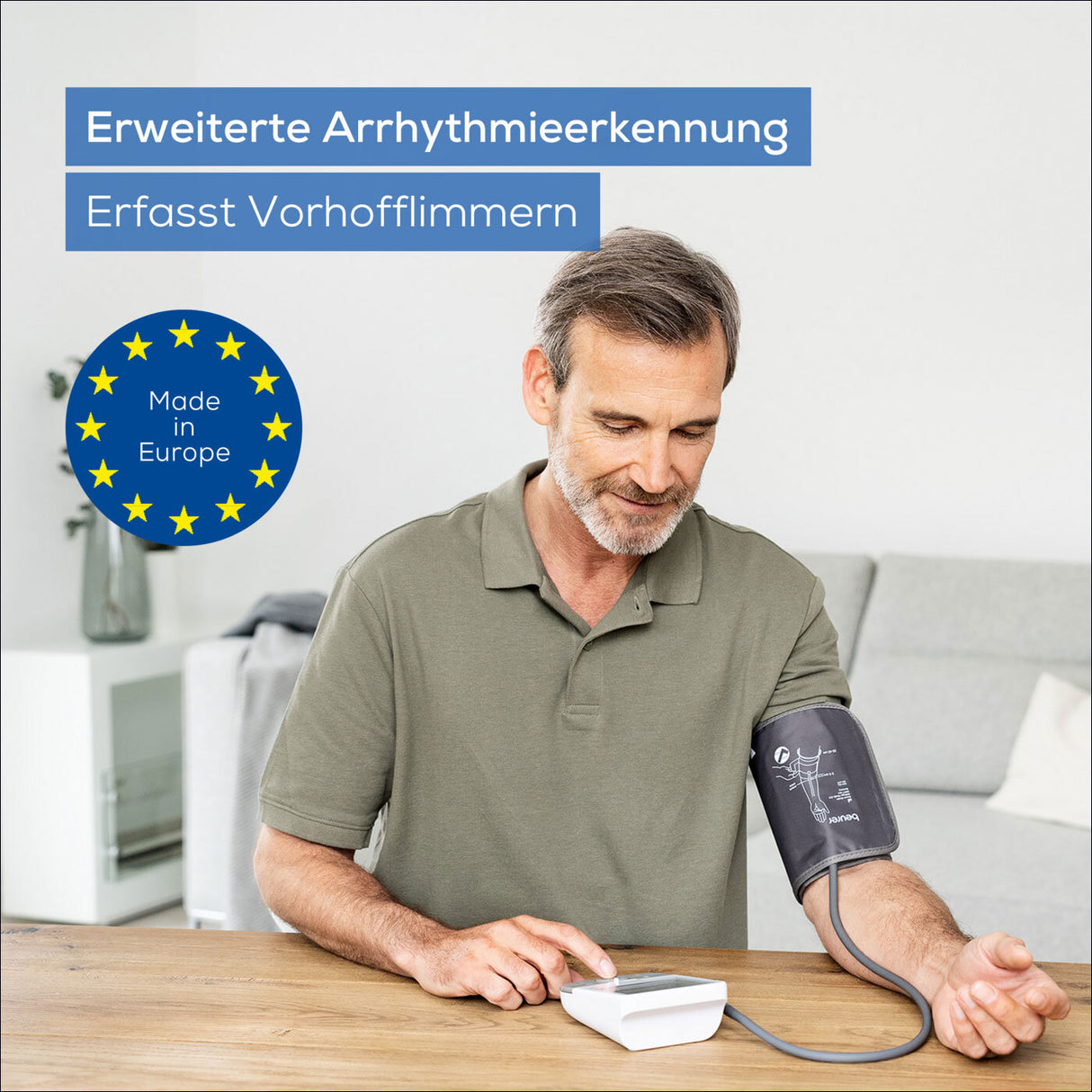 Älterer Mann mit Blutdruckmessgerät am Oberarm, sitzt an Tisch, im Hintergrund Sofa und Vase.
