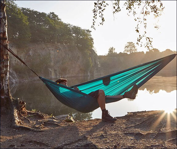 Person liegt in einer blauen Hängematte am Flussufer, Sonnenlicht und Bäume im Hintergrund.