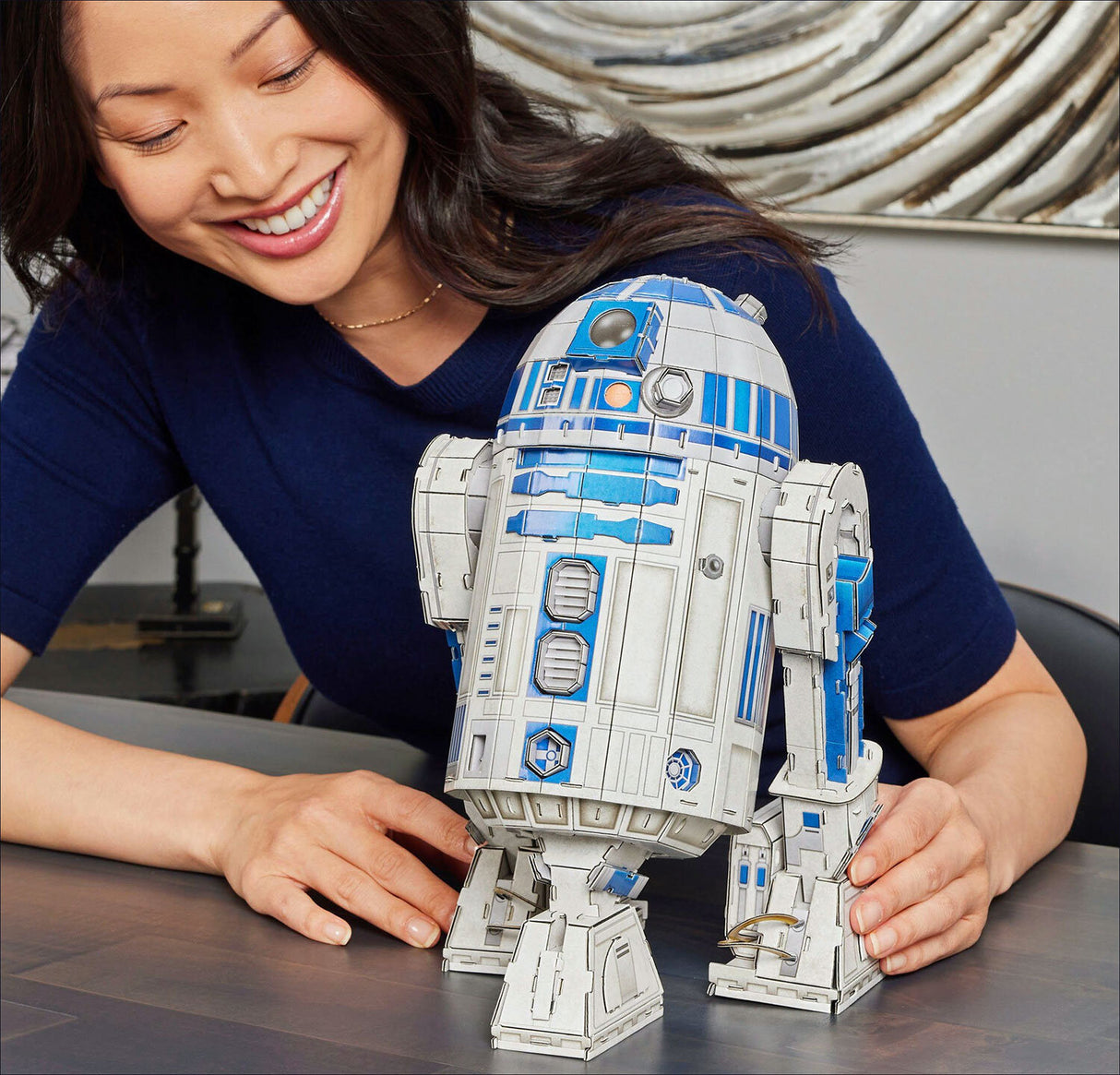 Frau lächelt neben einem R2-D2-Modell aus LEGO auf einem Tisch.