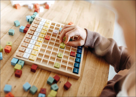 Hand eines Kindes beim Spielen mit einem numerischen Dominospiel auf einem Holztisch, umgeben von bunten Würfeln.