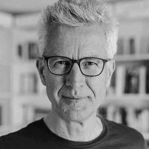 Schwarz-weiß-Bild eines Mannes mit grauem, zerzaustem Haar, Brille und dunklem Shirt vor einem Gebäude mit Fenstern.