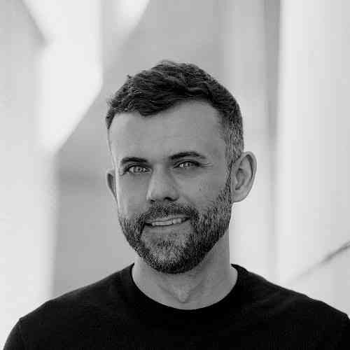 Das Bild zeigt einen Mann mit dunklem, kurzem Haar und Bart, in einem schwarzen Shirt, vor hellem Hintergrund.