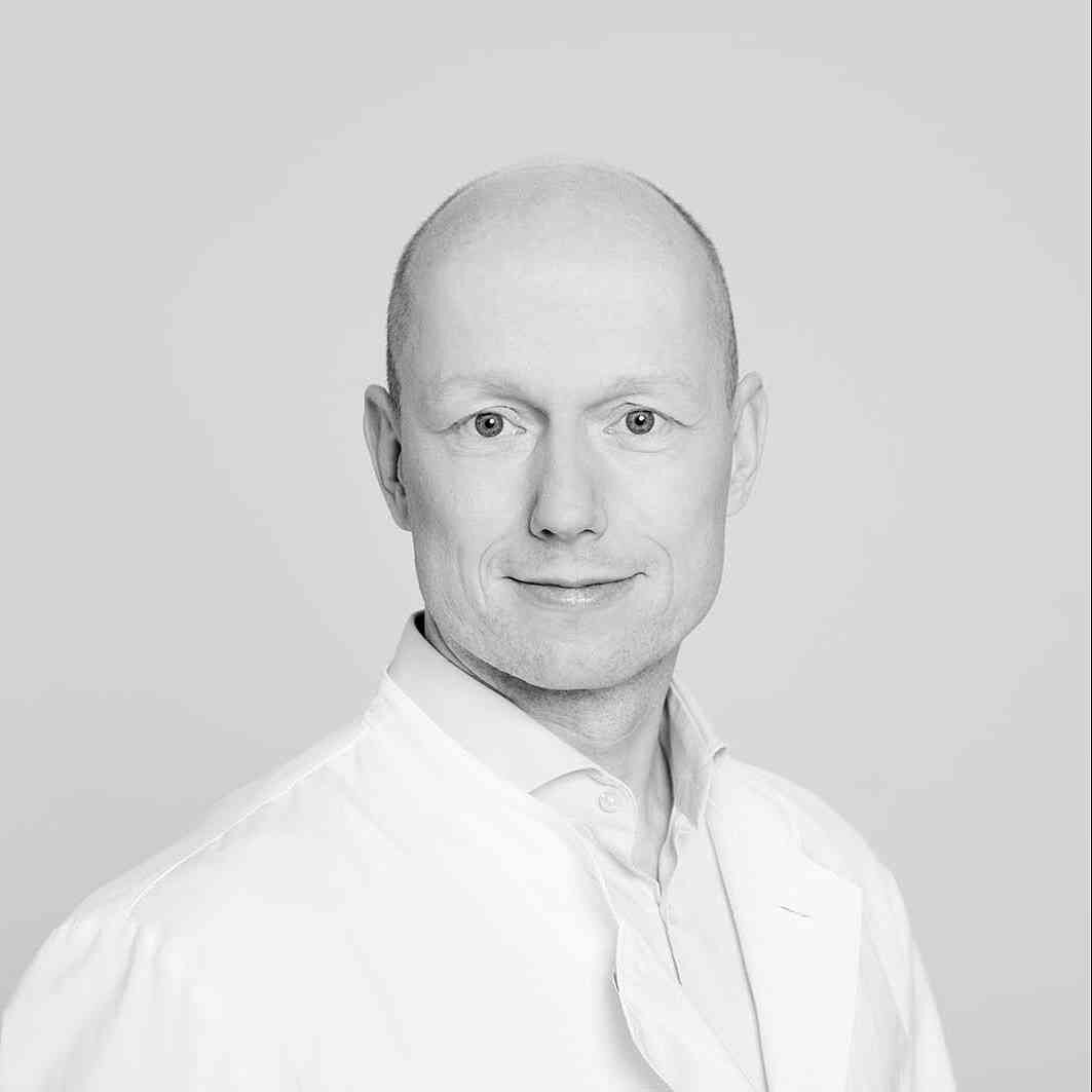 Schwarz-weiß-Porträt eines Mannes mit kurzen Haaren, weißem Hemd, lächelnd, vor hellem Hintergrund.