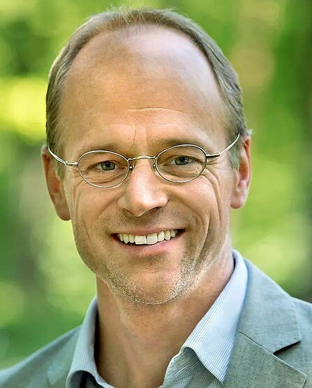 Mann mit Brille, lächelnd, im Freien mit grüner, unscharfer Natur im Hintergrund.