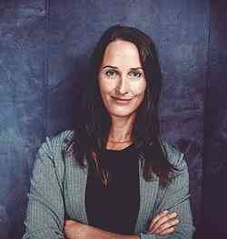 Frau mit dunklen Haaren, grauem Blazer, vor dunklem Hintergrund, lächelnd mit verschränkten Armen.