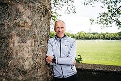Mann lehnt an Baum im Park, trägt Sportjacke, lächelt, grüne Wiese und Bäume im Hintergrund.