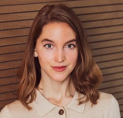 Junge Frau mit braunen, schulterlangen Haaren, trägt eine beige Jacke und steht vor einer Holzvertäfelung.