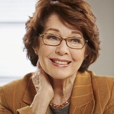 Lächelnde ältere Frau mit Brille, braune Haare, trägt eine braune Jacke und eine Perlenkette.