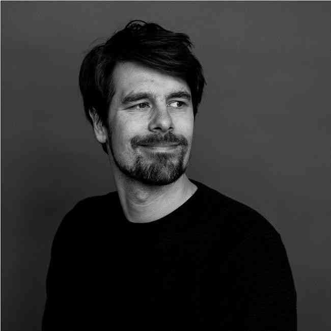 Schwarz-weiß-Porträt eines Mannes mit dunklem Haar, Bart und einem schwarzen Pullover, lächelnd und nach rechts blickend.