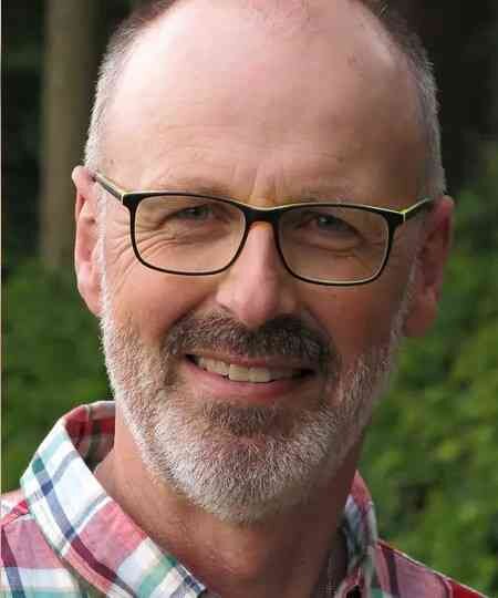 Nahaufnahme eines Mannes mit Brille, grauem Bart und kurzen Haaren, lächelnd vor grüner Natur.