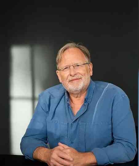 Ein älterer Mann mit Brille, grauem Haar und Bart, trägt ein blaues Hemd und sitzt vor dunklem Hintergrund.