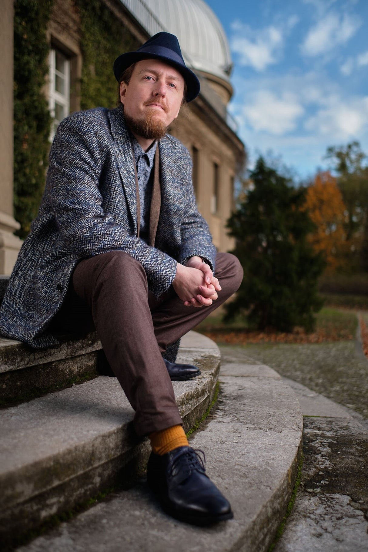 Junger Mann sitzt auf Steintreppe vor einem historischen Gebäude, trägt Hut, Jacke, Hose, Schuhe und bunte Socken.
