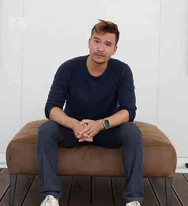 Junger Mann sitzt auf einer braunen Bank, trägt dunklen Pullover, Jeans und eine Armbanduhr, vor weißer Wand.