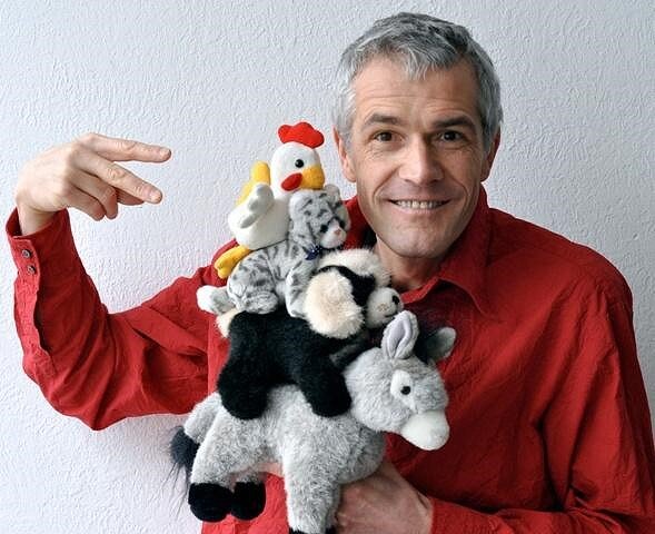 Ein Mann mit grauem Haar trägt ein rotes Hemd und hält eine Hand voller Plüschtiere, darunter eine Ente, Katzen