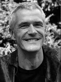 Schwarz-weiß-Bild eines lächelnden Mannes mit kurzen Haaren vor Bäumen und Pflanzen.