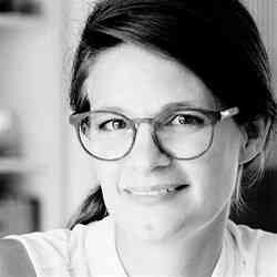 Frau mit Brille, dunklem Haar, lächelnd, in einem hellen Raum.