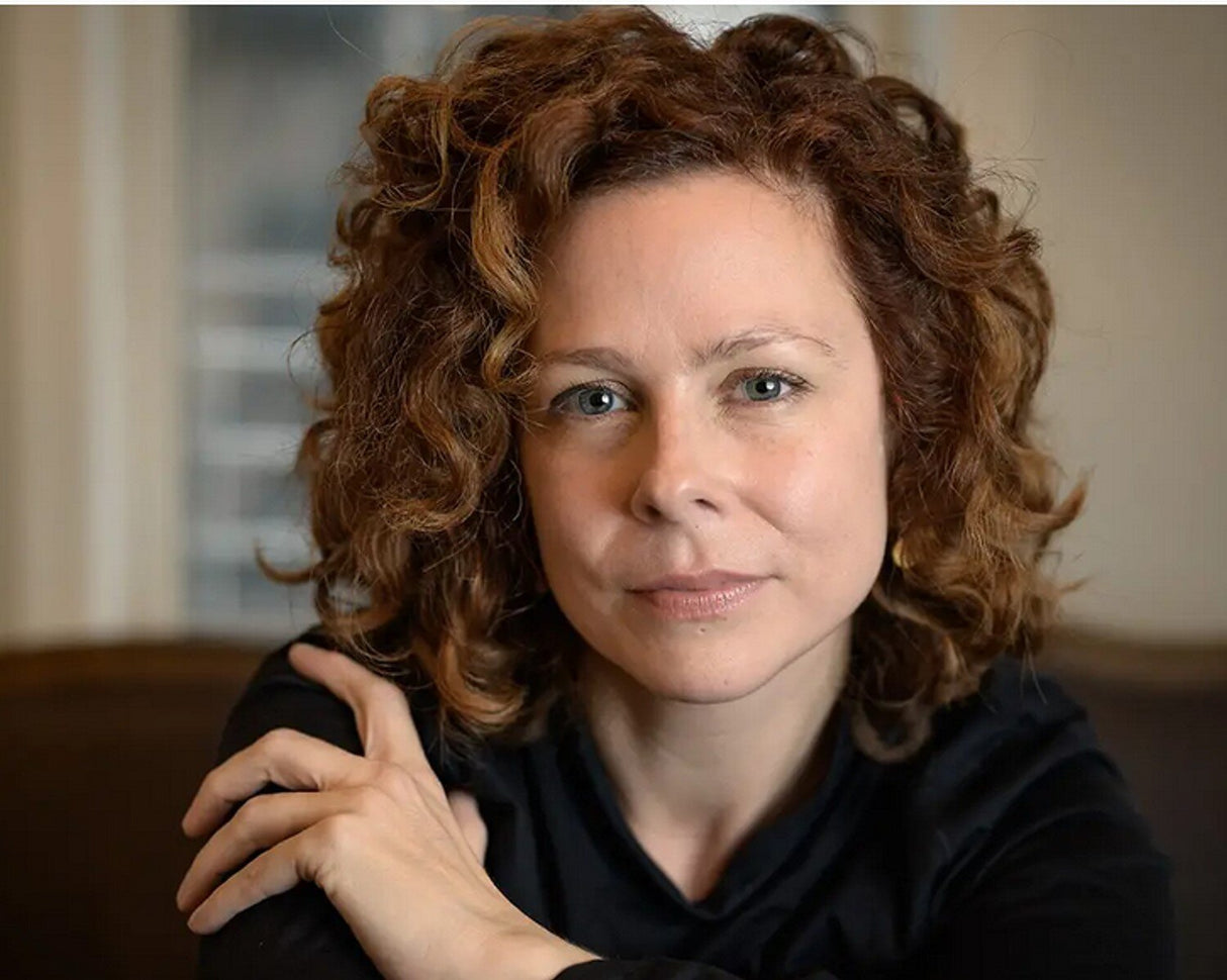 Porträt einer Frau mit lockigem, rotem Haar, blauen Augen und schwarzem Oberteil, die direkt in die Kamera schaut.