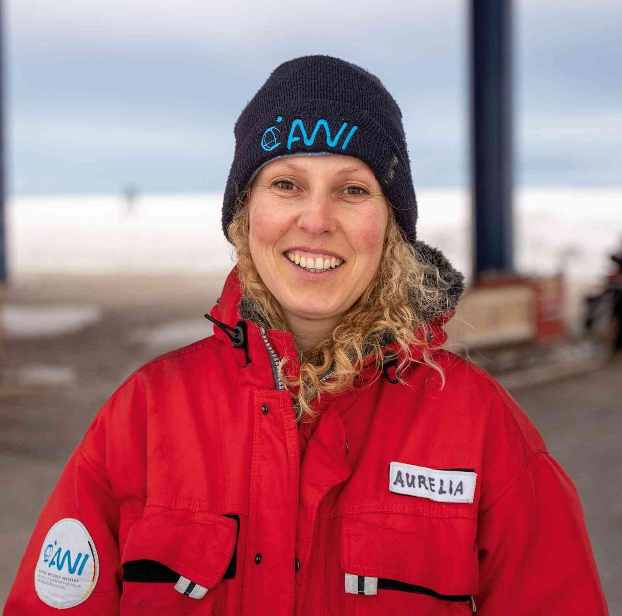 Frau mit roter Jacke, schwarzer Mütze mit blauer Aufschrift, Namensschild "Aurelia", lächelnd im Freien bei bewölktem Himmel.