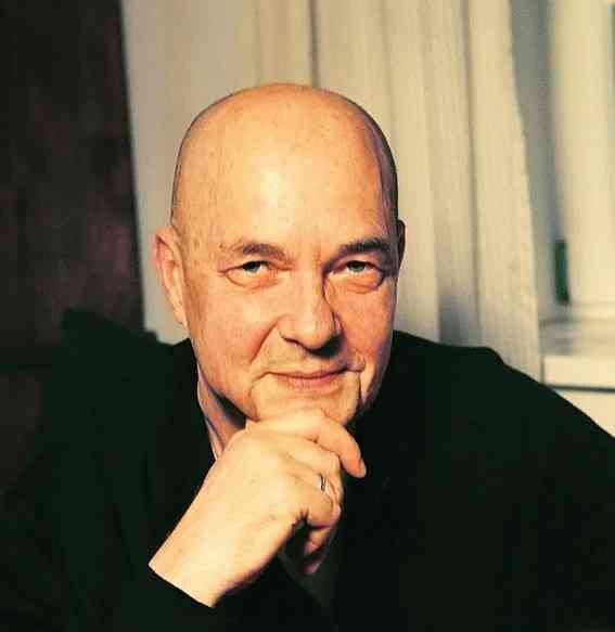 Baldköpfiger Mann mit Hand an Kinn, leicht lächelnd, im Hintergrund Vorhänge.