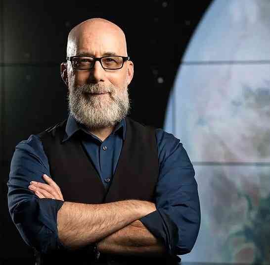 Mann mit Brille, grauem Bart, schwarzen Hemd und Weste, mit verschränkten Armen vor dunklem Hintergrund.