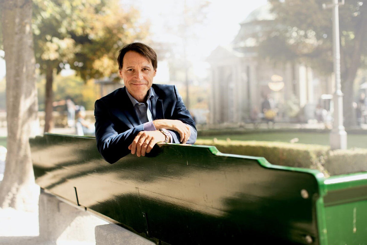 Mann sitzt an einer Parkbank im Freien, im Hintergrund Bäume und Gebäude, bei Sonnenlicht.