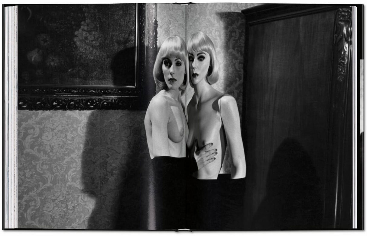 Zwei Frauen mit kurzen blondem Haar, nackt, stehen eng beieinander vor einer Wand mit Tapete und einem großen Rahmen.