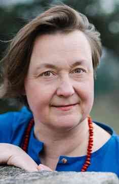 Nahaufnahme eines älteren Frauengesichts mit braunen Haaren, blauer Kleidung und roter Halskette.