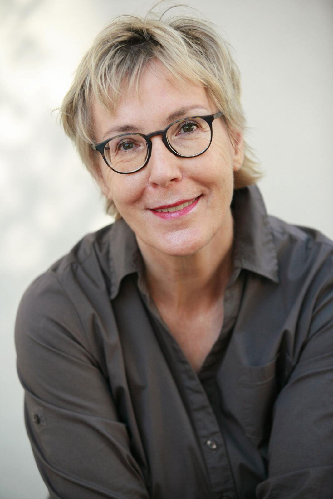 Porträt einer lächelnden Frau mit kurzen, blonden Haaren, Brille und grauem Hemd vor hellem Hintergrund.