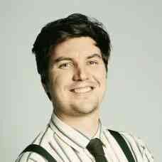 Ein Mann mit dunklem Haar, trägt ein gestreiftes Hemd und eine schwarze Krawatte, lächelt und zwinkert.