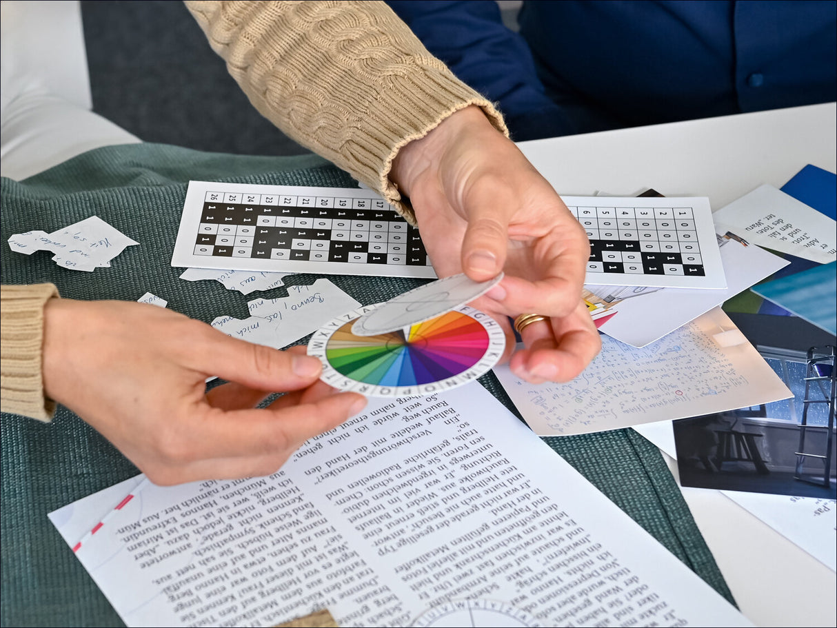 Person hält Farbkreis und Münze über Tisch mit Papieren, Notizen und Farbtafeln.