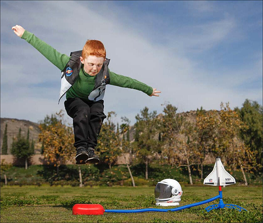 Kind mit roten Haaren springt in der Luft, vor einem Modellraketen-Startset im Freien.
