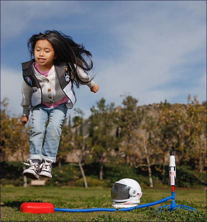 Mädchen springt in einem Park, neben einem Raumfahrt-Set mit Helm, Rakete und Startpad.