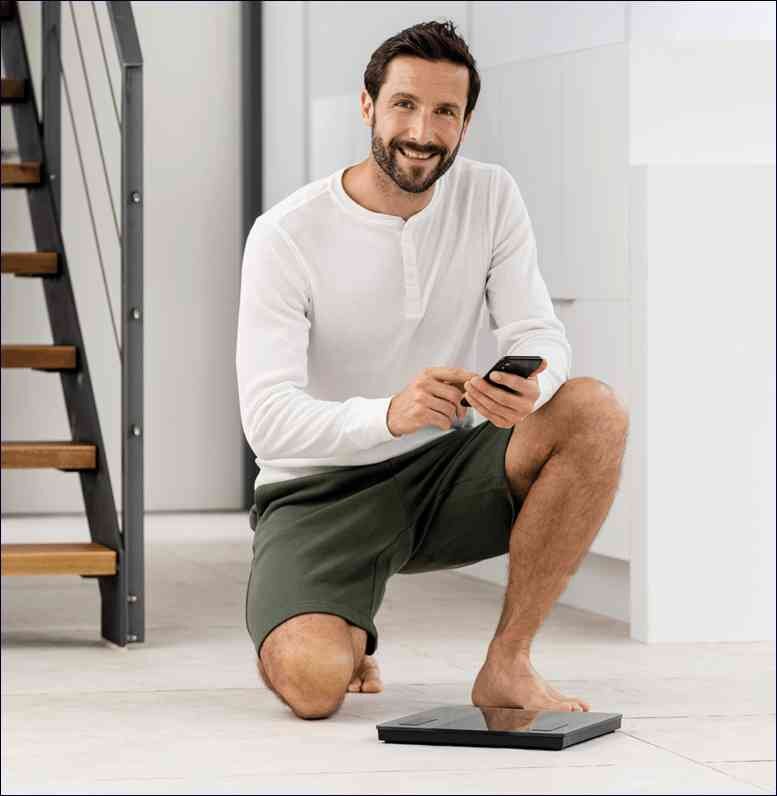 Mann steht auf Waage, hält Smartphone, lächelt, im modernen Zuhause.