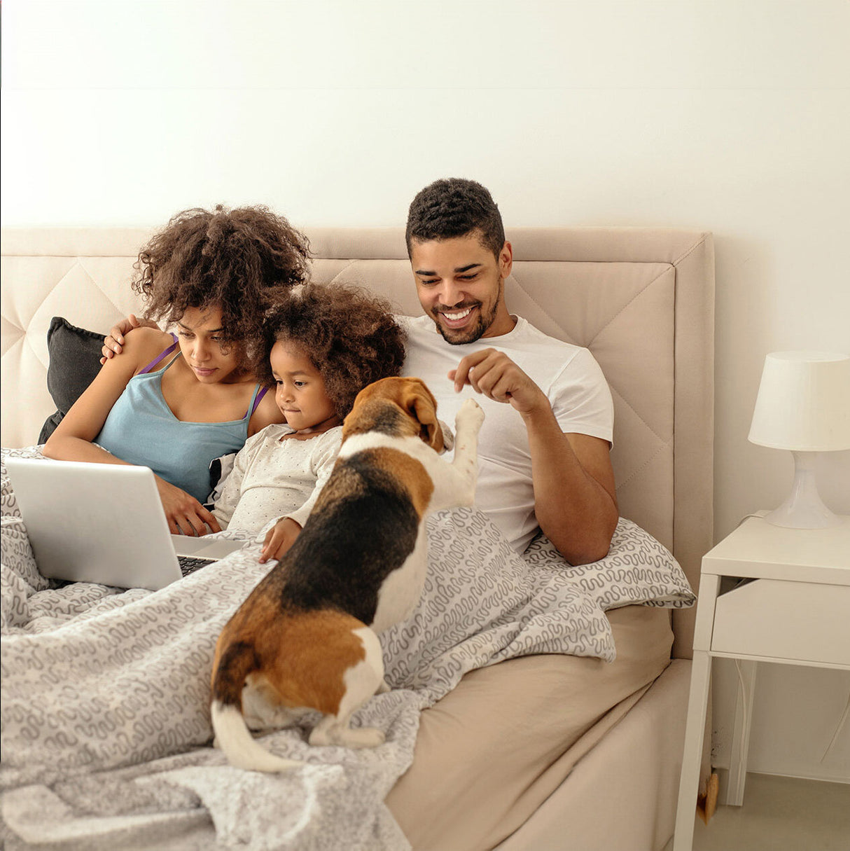 Familie mit zwei Kindern und Hund sitzt im Bett, schaut auf Laptop, lächelt und spielt mit dem Hund.