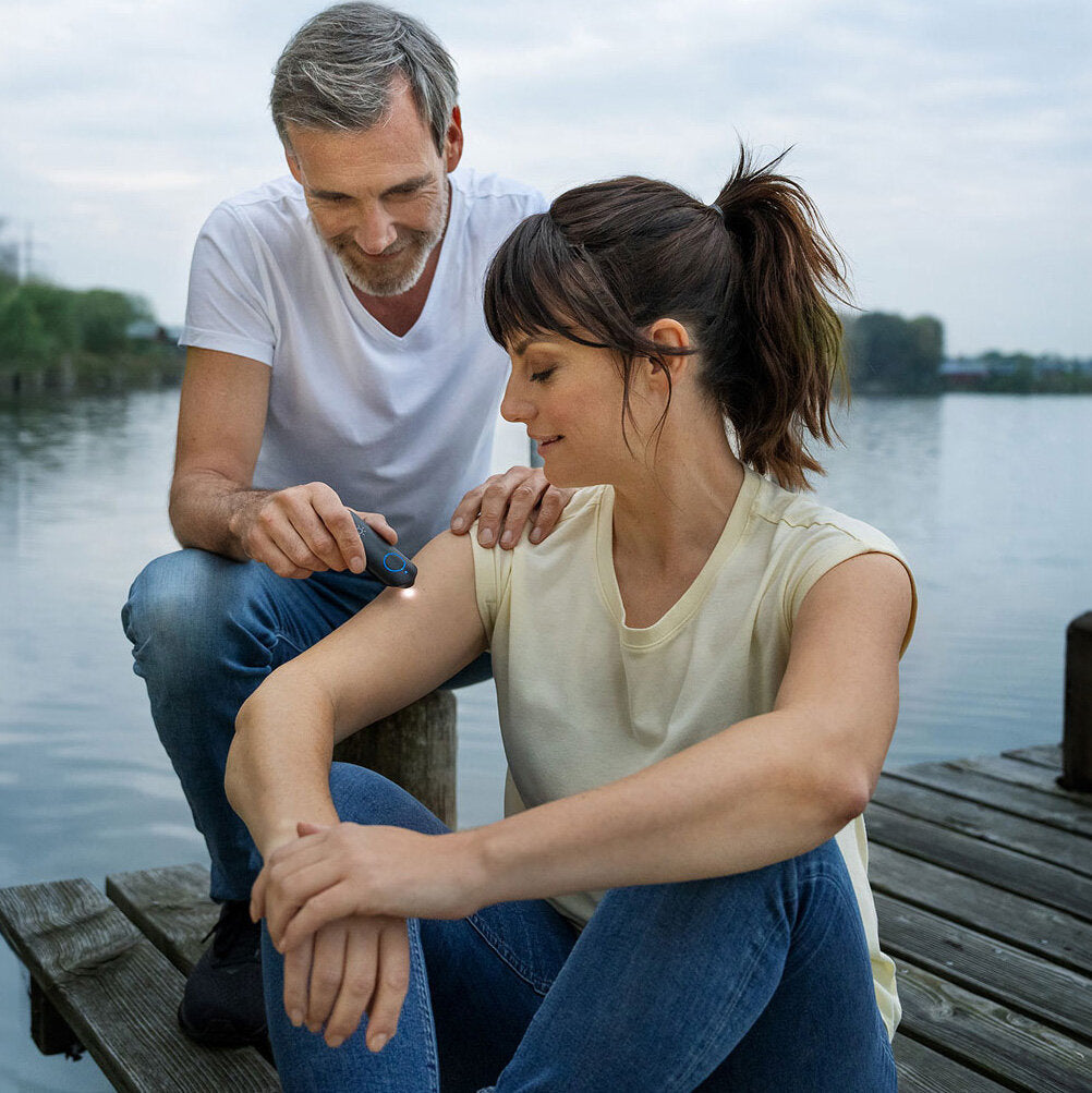 Mann misst die Temperatur am Oberarm einer sitzenden Frau mit einem Infrarot-Thermometer am See.