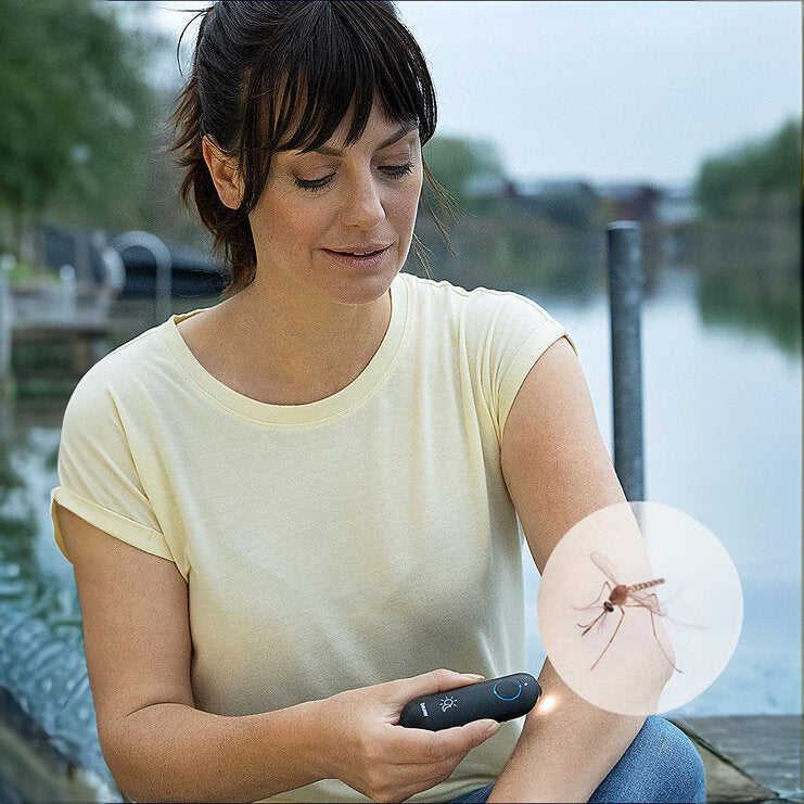 Frau hält Insektenschutzmittel vor den Arm, mit Wasser im Hintergrund.