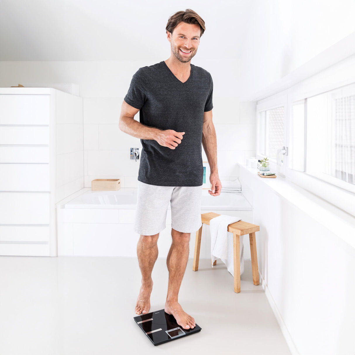 Mann steht auf einer Waage im Badezimmer, trägt T-Shirt und Shorts, lächelt.
