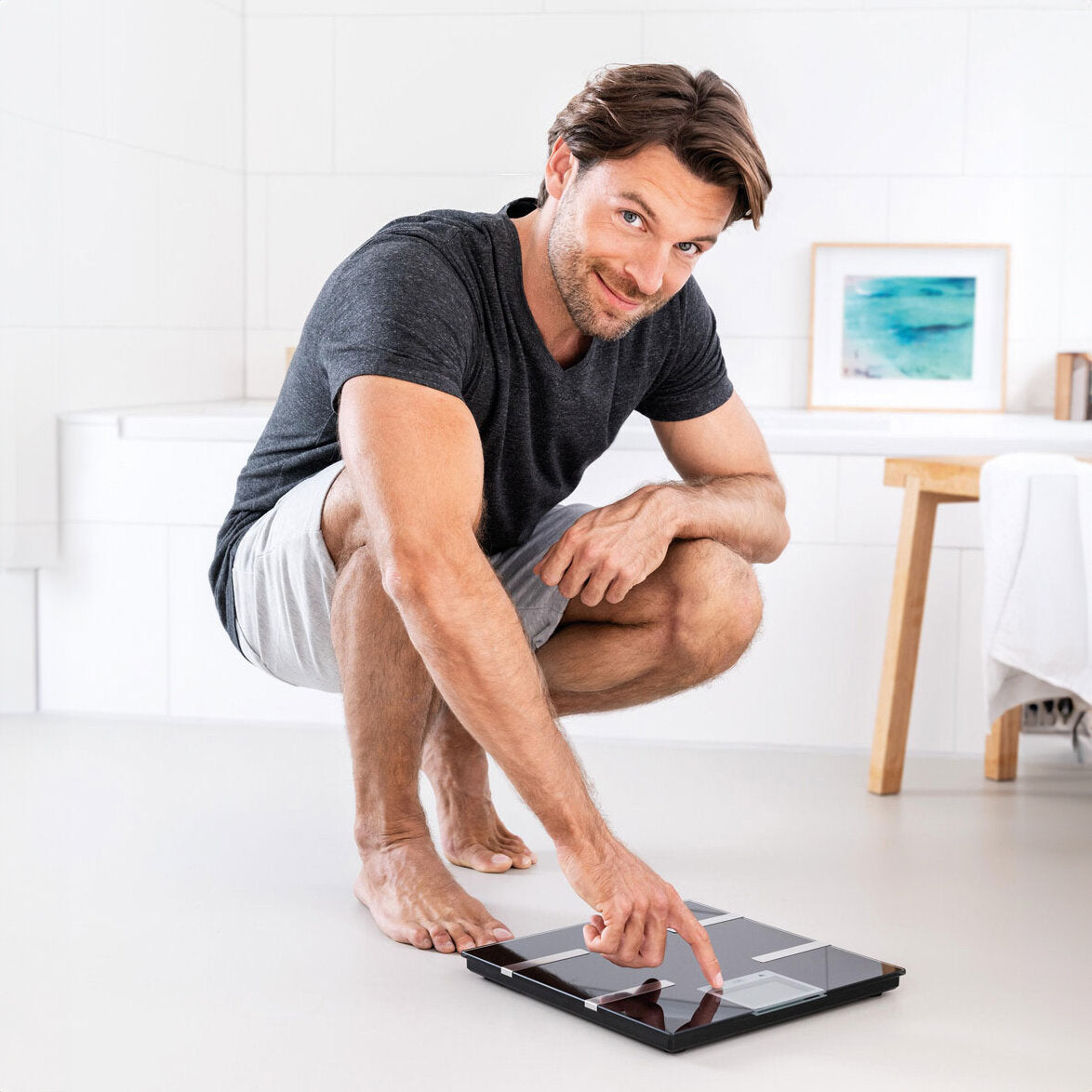 Mann im schwarzen T-Shirt und Shorts steht auf Zehenspitzen, zeigt auf eine Waage auf dem Boden.