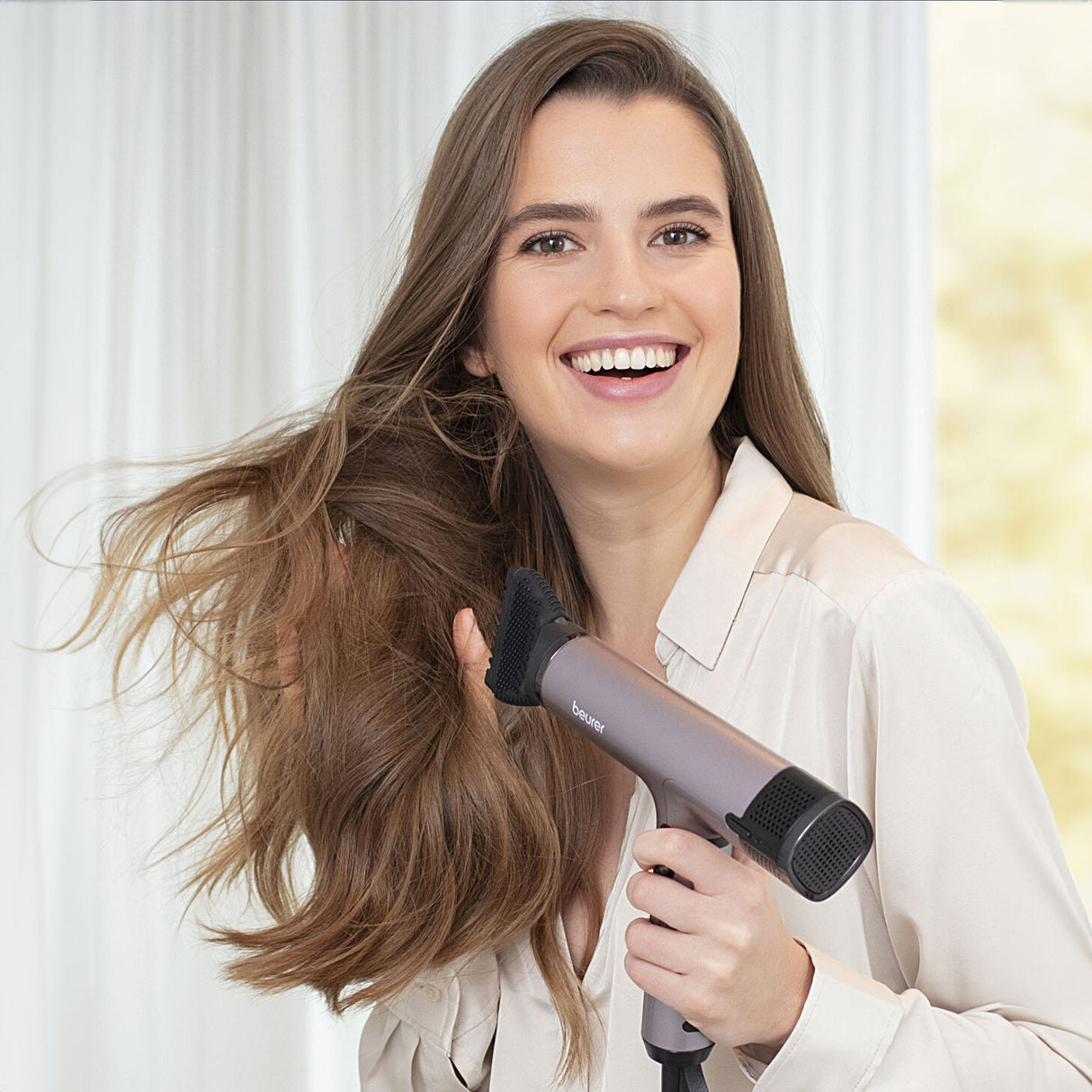 Junge Frau trocknet ihr langes, braunes Haar mit einem Föhn, lächelt und trägt eine weiße Bluse.