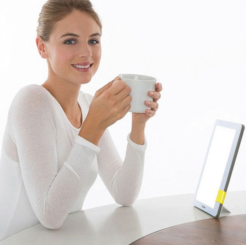 Junge Frau mit hellbraunen Haaren, lächelnd, hält weißen Kaffeebecher, sitzt an einem weißen Tisch, neben einem Tablet.
